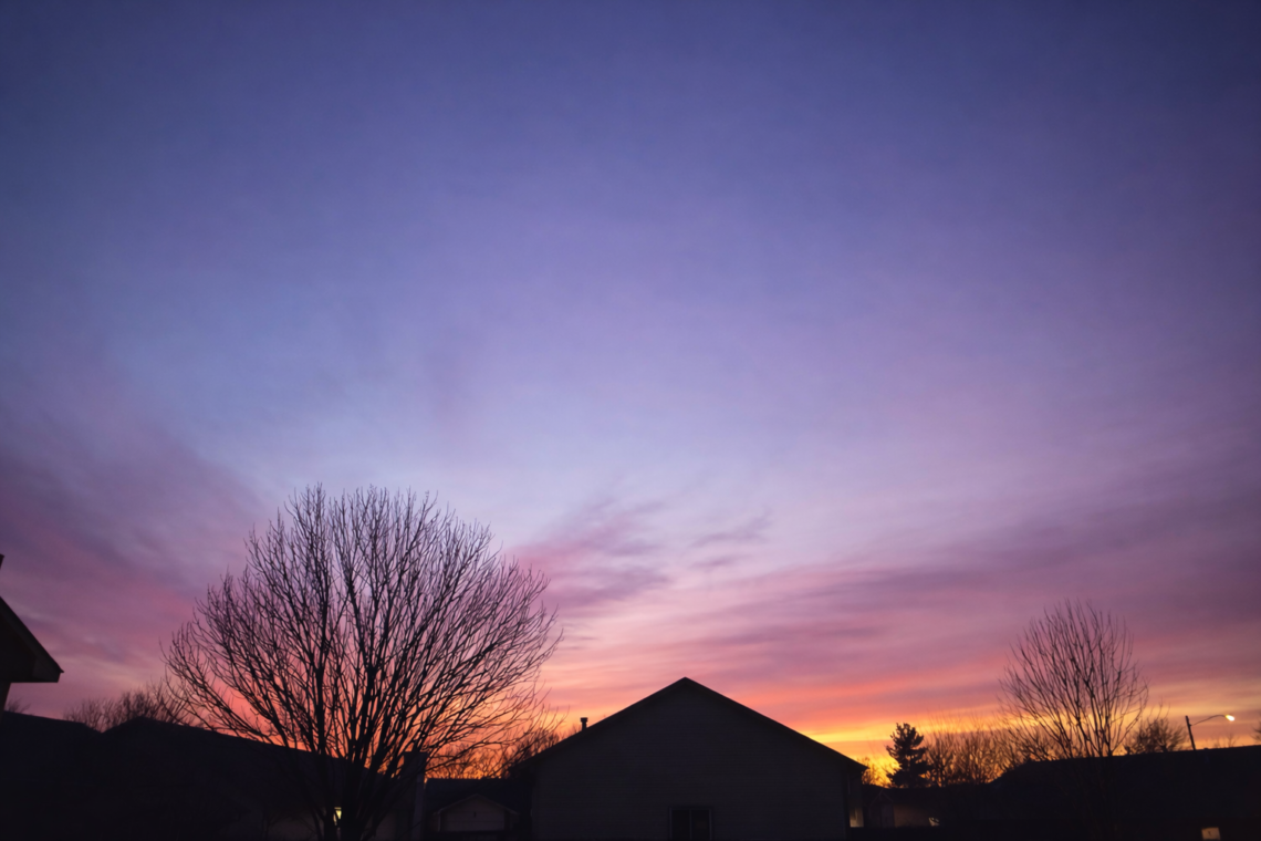 Soft winter sunset with purple, pink, and golden sky over silhouetted trees and rooftops